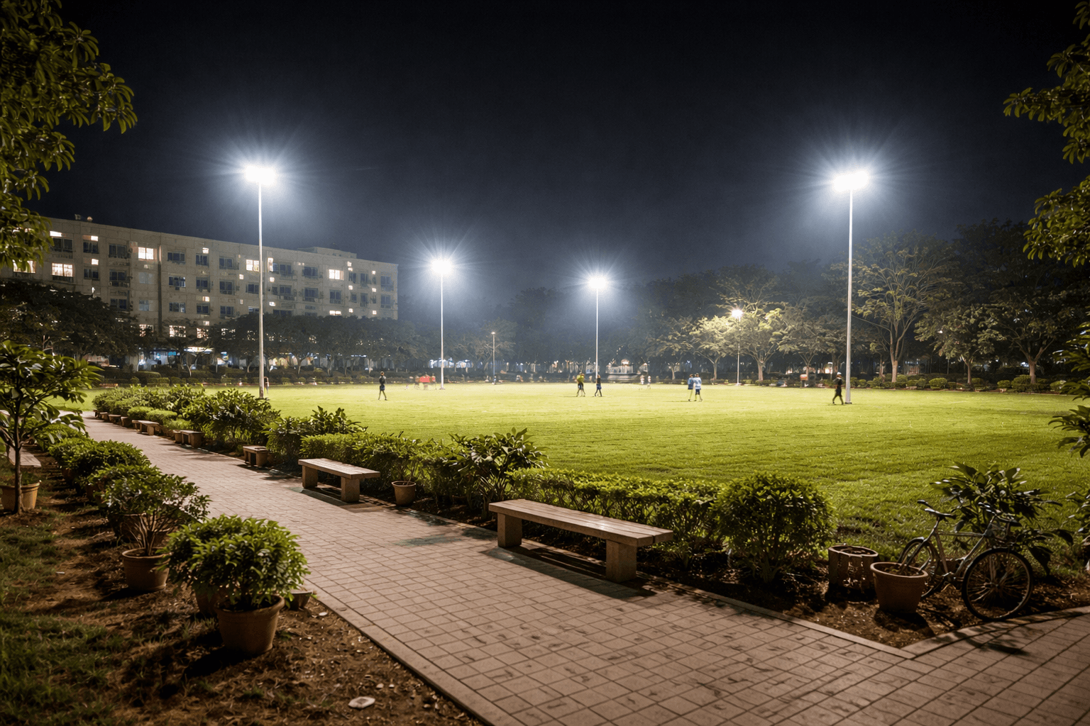 People playing on a brightly lit green field at night, seen from a paved walkway.